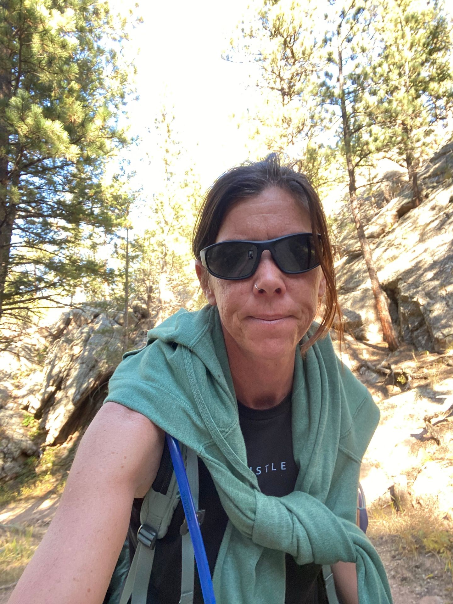 Person wearing sunglasses and a green sweater tied around the shoulders, standing in a forested area.