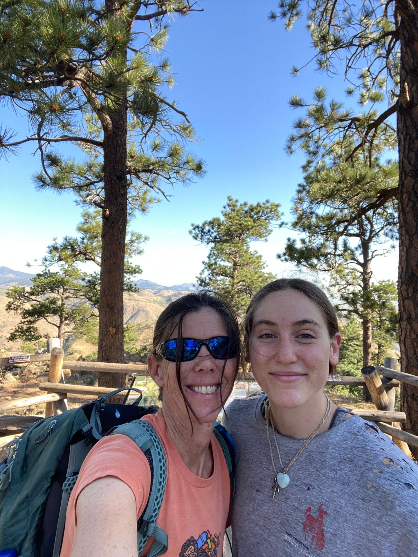 Two people smiling outdoors with pine trees and mountains in the background.