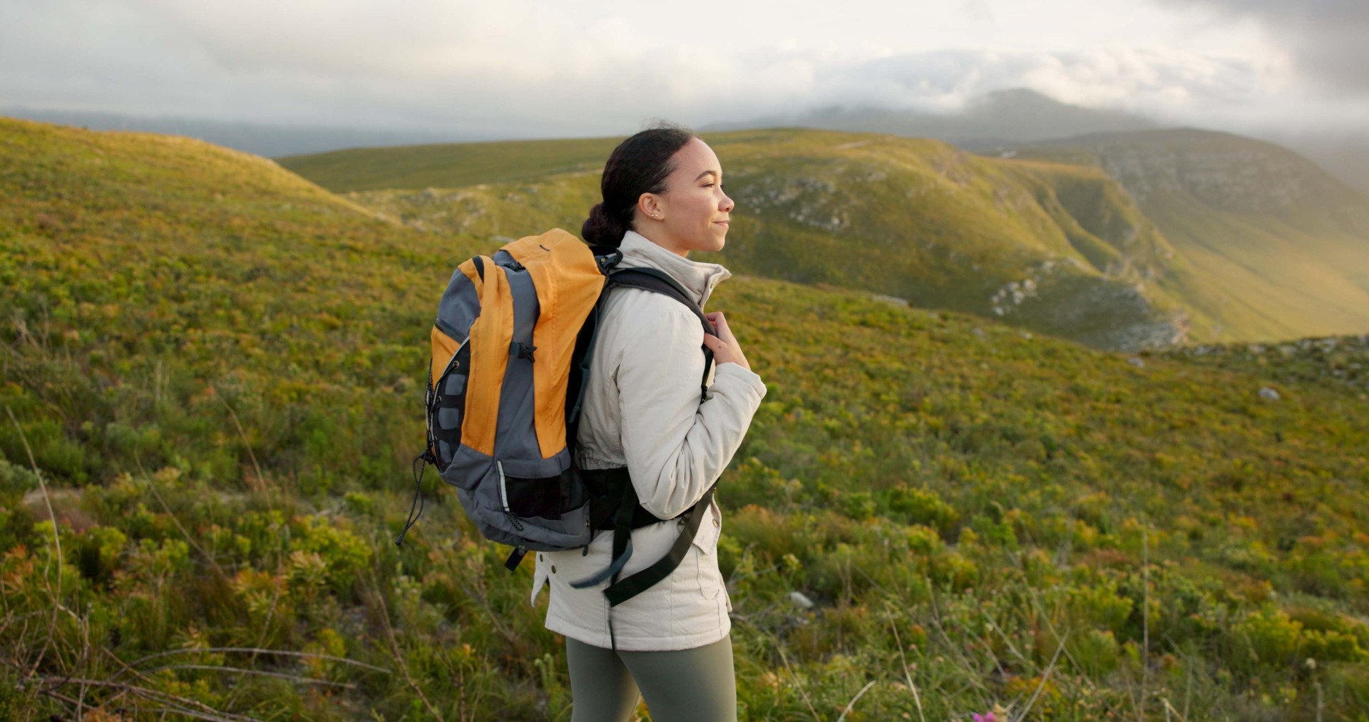 Woman, hiking on mountains and breathing fresh air for outdoor wellness, fitness and health in nature. Happy young person in wind with backpack and trekking on a hill for adventure, travel or journey